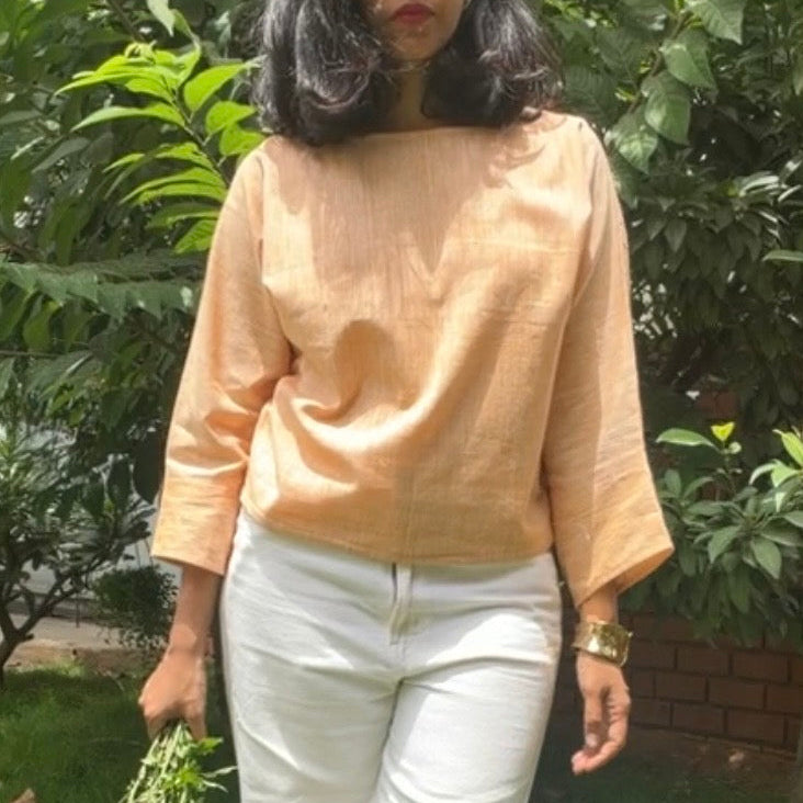 A woman standing outdoors on a grassy lawn in front of green bushes, wearing a peach-colored khadi cotton linen three-quarter sleeve top and white trousers, holding a small bunch of purple flowers.